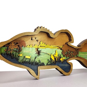 Op de afbeelding: Een gelaagd houten kunstwerk in de vorm van een vis, dat een visscène uitbeeldt. Het kunstwerk toont een meer met een visser, eenden, bomen en vogels tegen een zonsondergang. Het stuk is gemaakt van natuurlijke houttinten met geschilderde details.
