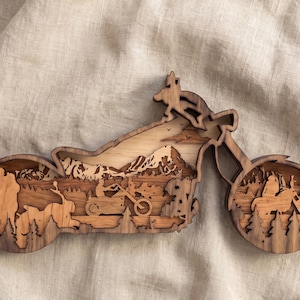 May include: A multi-layered wooden wall art piece shaped like a motorcycle silhouette. The design features intricate cutouts of a person riding a motorcycle, a standing figure, a forest landscape, and mountain peaks, all crafted from various wood tones.