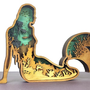 May include: Wooden mermaid sculpture with layered design. The sculpture features a mermaid silhouette with a detailed underwater scene, including a shipwreck, coral, and fish. The layers are stained in shades of brown, green, and yellow.
