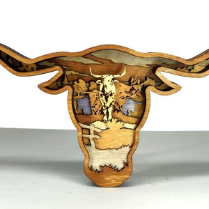 May include: A layered wooden sculpture of a longhorn steer head. The artwork features a detailed landscape scene within the steer's head, with various shades of brown and tan. The horns extend outward, creating a unique decorative piece.