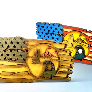 May include: Two wooden American flag-themed wall art pieces. One features a natural wood finish with a circle design of an eagle, mountains, and trees. The other has a blue star field and red stripes, with a similar design. Both are layered and detailed.