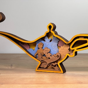 May include: A layered wooden sculpture of a genie lamp featuring characters from a popular animated film. The lamp is primarily brown with yellow and orange accents, and the interior depicts a scene with figures and a blue sky.