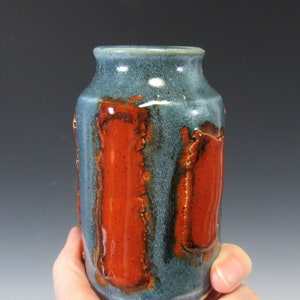 Può includere: Un piccolo vaso in ceramica blu con una superficie testurizzata e accenti rossi, marroni e neri. Il vaso è fatto a mano e ha un aspetto rustico unico.