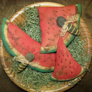 May include: Three handmade watermelon slices with green edges and brown stitching. Each slice has a black and yellow bee appliqué. The watermelons are displayed in a wooden bowl with green moss.