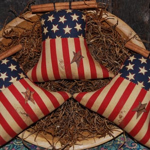 May include: Three patriotic star-spangled and striped fabric decorations in a wooden bowl with cinnamon sticks. The decorations are red, white, and blue, with a distressed, vintage look. The bowl is filled with dried grass.