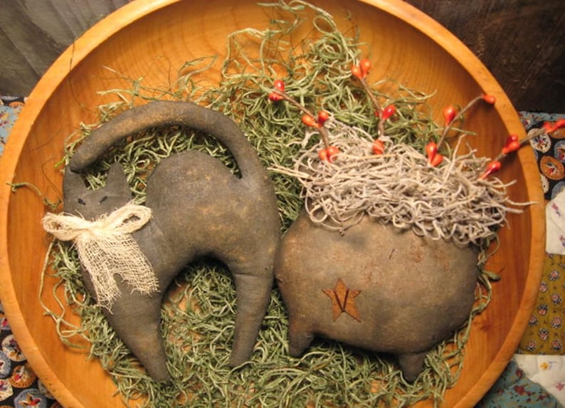Op de afbeelding: Een rustieke houten kom bevat een zwart kattenbeeldje met een jute strik en een ketelvormige decoratie. De ketel heeft een ster en is gevuld met gedroogd mos en rode bessen, wat een Halloween-thema oproept.