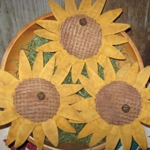 May include: Three handcrafted sunflowers with yellow petals and brown centers are arranged in a wooden bowl filled with green filler. The sunflowers are a rustic, country-style decoration.