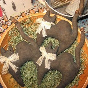 Op de afbeelding: Drie donkerbruine stoffen kattenfiguren met gestikte gelaatstrekken en staarten. Elke kat draagt een cr&egrave;mekleurige, gerafelde stoffen strik om de nek. Aan de uiteinden van de staarten van twee kattenfiguren zijn kleine metalen belletjes bevestigd.
