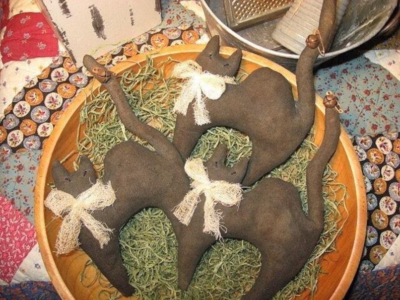 Op de afbeelding: Drie donkerbruine stoffen kattenfiguren met gestikte gelaatstrekken en staarten. Elke kat draagt een cr&egrave;mekleurige, gerafelde stoffen strik om de nek. Aan de uiteinden van de staarten van twee kattenfiguren zijn kleine metalen belletjes bevestigd.