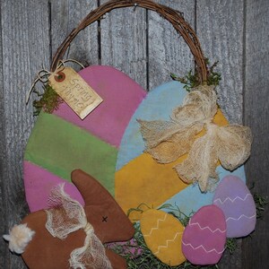 Puede incluir: Adorno de pared con temática de Pascua con asa tejida. El diseño presenta grandes formas de huevo coloridas en rosa, verde y azul, junto con un conejito marrón y huevos decorativos más pequeños. Una etiqueta dice "Spring Thyme."
