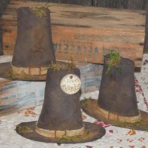 May include: Three brown pilgrim hats with wide brims and tan bands. Each hat is decorated with green moss and has a weathered look. One hat has a tag that says "Give Thanks".
