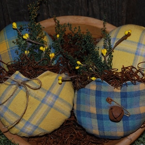 May include: Two fabric Easter eggs decorated with a blue and yellow plaid pattern. The eggs are nestled in a brown nest with yellow sprigs of greenery.