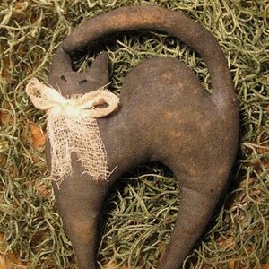 Op de afbeelding: Een donkerbruin, opgezette kattenfiguur met een gebogen staart en een cr&egrave;mekleurige jute strik om de nek. De kat staat tegen een achtergrond van gedroogd groen mos, wat een rustieke, landelijke esthetiek cre&euml;ert.