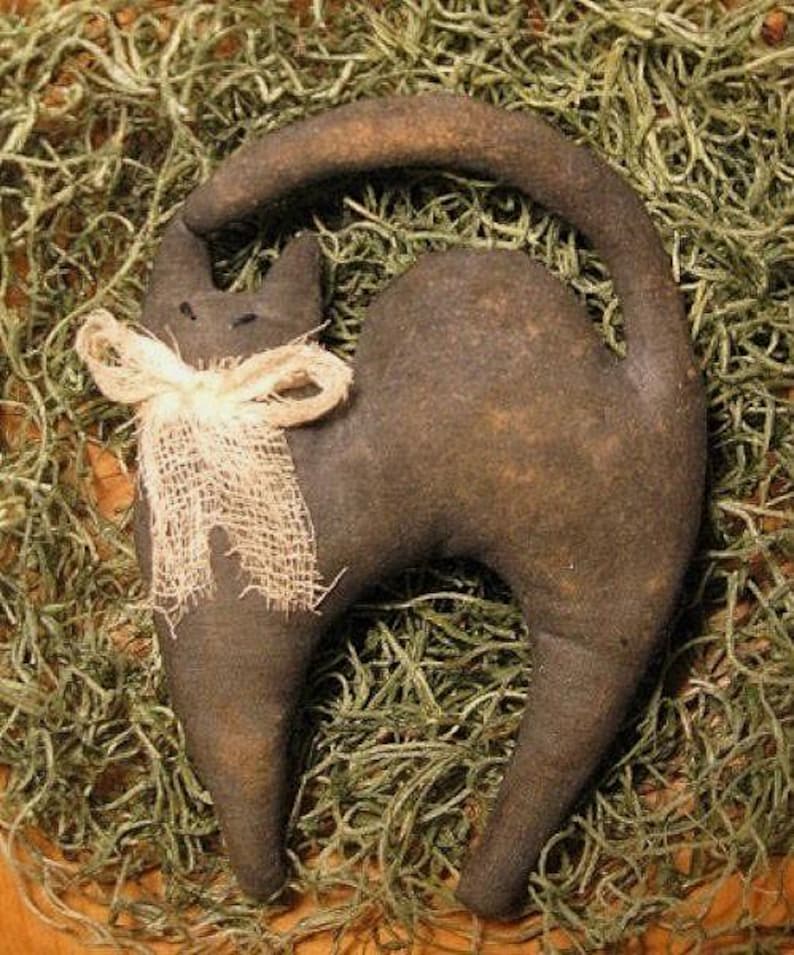 Op de afbeelding: Een donkerbruin, opgezette kattenfiguur met een gebogen staart en een cr&egrave;mekleurige jute strik om de nek. De kat staat tegen een achtergrond van gedroogd groen mos, wat een rustieke, landelijke esthetiek cre&euml;ert.