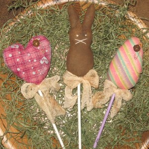 Op de afbeelding: Drie handgemaakte paasversieringen op stokjes. Een roze en wit hart, een bruin konijn en een regenboogkleurig gestreept ei. Elke versiering heeft een strik van jute en een bel.