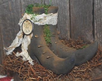 May include: A pair of dark brown fabric boots with pointed toes, decorated with green moss, white frayed fabric strips, and metal safety pins. A round tag attached to the side features the handwritten text Boots for Sale.