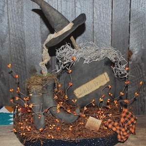 May include: A Halloween display with a witch's hat, cauldron, and boots. The cauldron has a label that reads "Witch's Brew". The display is decorated with orange berries and a black and orange plaid accent.