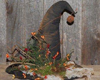 May include: A black witch's hat with a pointed crown, decorated with sprigs of greenery, red berries, and a small bell. The hat is sitting on a rustic wooden surface.