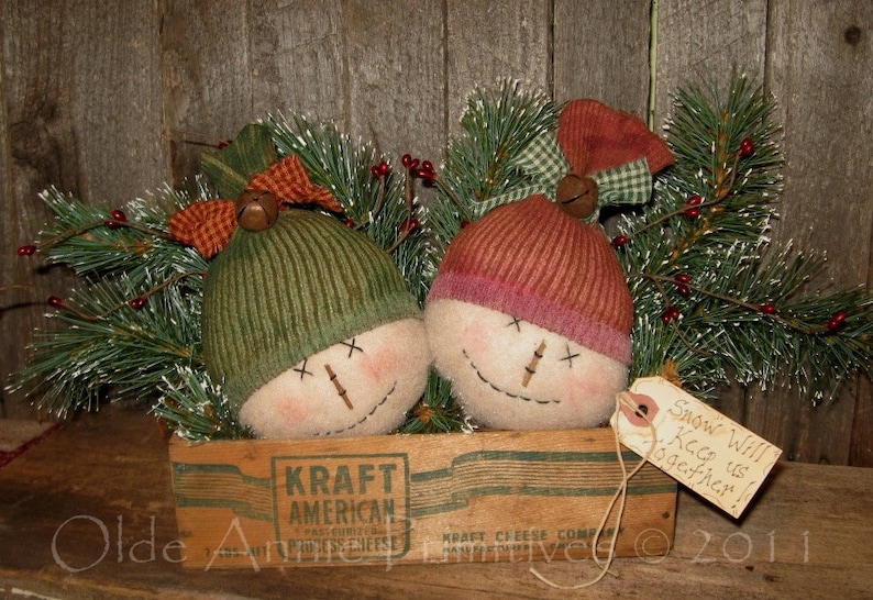 May include: Two handmade fabric snowmen with red and green hats and bells, sitting in a vintage Kraft American Process Cheese box with a tag that reads "Snow Will Keep us Together!"