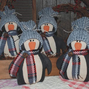 Op de afbeelding: Vijf handgemaakte pinguïnornamenten. Elke pinguïn heeft een grijze gebreide muts, een rood, wit en blauw geruite sjaal en een kleine bel. De pinguïns zijn zwart en wit met oranje snavels en worden tentoongesteld in een houten kist.