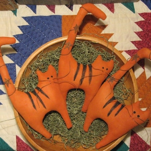 May include: Three orange fabric cats with black stripes are sitting in a wooden bowl filled with green moss. The cats have long, thin tails and are posed in a playful manner.