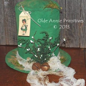 Puede incluir: Un sombrero de copa verde con ala ancha, decorado con una ramita de vegetación, bayas blancas y dos campanas de bronce. Se adjunta una etiqueta con una ilustración vintage de una niña con un vestido verde. El sombrero está adornado con tela color crema.