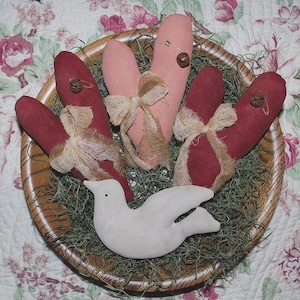 Könnte beinhalten: Ein Holzkörbchen gefüllt mit dekorativen Herzen und einer weißen Taube. Die Herzen sind in Rot- und Rosatönen gehalten und mit Schleifen aus Jute und kleinen Glöckchen verziert. Die Taube ist aus einem weichen, cremefarbenen Stoff.