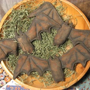 May include: A wooden bowl filled with dried green moss and several brown felt bats. The bats have detailed wings and are arranged in a circular pattern, creating a Halloween or autumn theme.