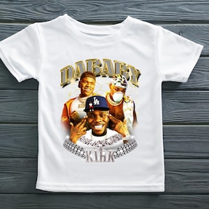 May include: White t-shirt with a graphic print of three men wearing gold chains and hats. The text "DaBaby Kirk" is printed on the shirt.