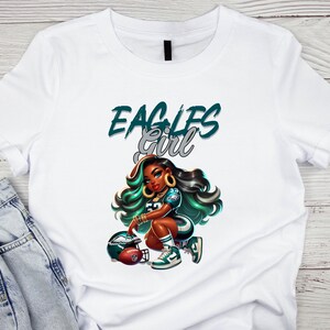 Pu&ograve; includere: T-shirt bianca con un'illustrazione a cartoni animati di una donna in uniforme da football, con la scritta "EAGLES Girl" in verde acqua e argento. La donna &egrave; inginocchiata accanto a un casco da football.