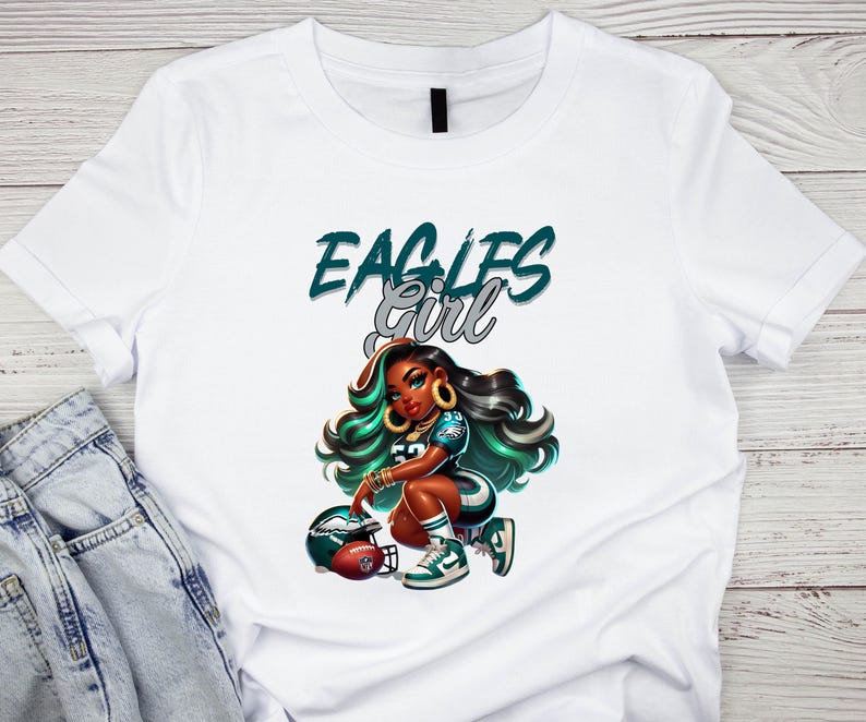 Pu&ograve; includere: T-shirt bianca con un'illustrazione a cartoni animati di una donna in uniforme da football, con la scritta "EAGLES Girl" in verde acqua e argento. La donna &egrave; inginocchiata accanto a un casco da football.
