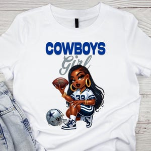 Pu&ograve; includere: T-shirt bianca con un'illustrazione a cartoni animati di una donna inginocchiata che tiene un pallone da football. La scritta "COWBOYS Girl" &egrave; in blu e grigio. &Egrave; visibile anche un casco da football.