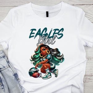 Pu&ograve; includere: T-shirt bianca con un'illustrazione a cartoni animati di una donna in uniforme da football, con la scritta "EAGLES GIRL" in verde acqua e argento. La donna &egrave; inginocchiata accanto a un pallone da football e a un casco, con dettagli verdi e neri.