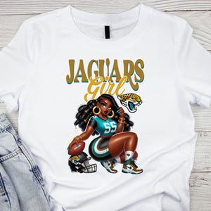Pu&ograve; includere: T-shirt bianca con un'illustrazione a cartoni animati di una donna in uniforme da football turchese e nera, numero 55. Il testo "Jaguars Girl" &egrave; sopra l'illustrazione, insieme a un logo di giaguaro e un pallone da football.