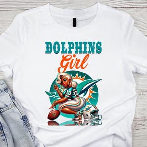 Pu&ograve; includere: T-shirt bianco con grafica di una donna in uniforme da football, la scritta "DOLPHINS Girl" e il logo dei Miami Dolphins. Il design include un pallone da football e un casco. La maglietta &egrave; abbinata a jeans blu.