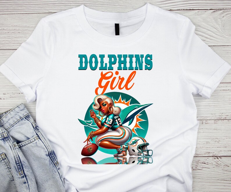 Pu&ograve; includere: T-shirt bianco con grafica di una donna in uniforme da football, la scritta "DOLPHINS Girl" e il logo dei Miami Dolphins. Il design include un pallone da football e un casco. La maglietta &egrave; abbinata a jeans blu.
