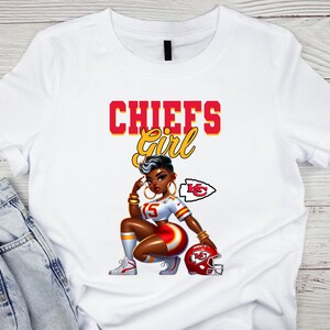 Pu&ograve; includere: T-shirt bianca con grafica di una donna in uniforme da football dei Chiefs, con la scritta "CHIEFS Girl" in rosso e giallo. La donna indossa un'uniforme rossa e bianca, gioielli dorati e tiene in mano un casco rosso.