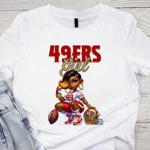 Pu&ograve; includere: T-shirt bianco con un'illustrazione a cartoni animati di una donna in uniforme da football. Il testo "49ERS Girl" &egrave; sopra l'illustrazione in rosso e oro. La donna indossa un'uniforme rossa e bianca, con un pallone da football e un casco dorato.