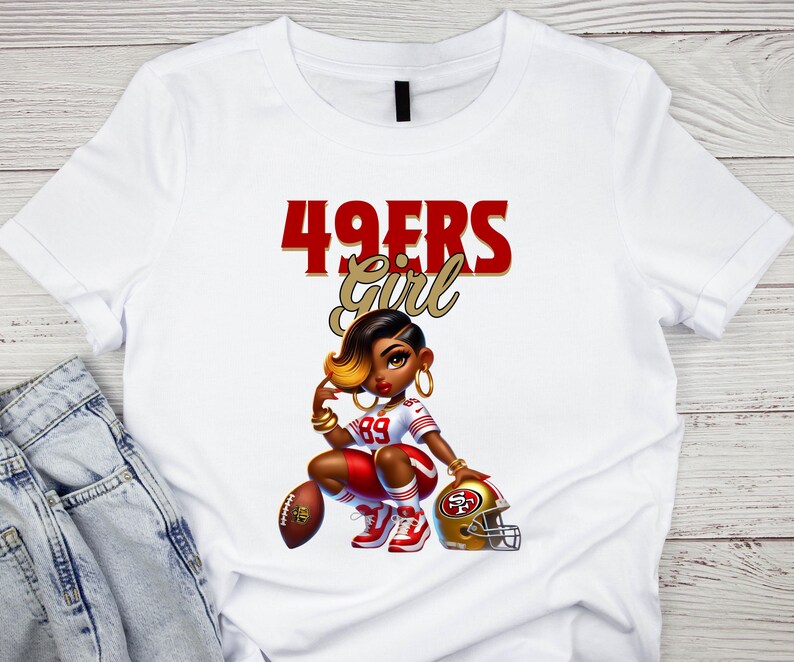 Pu&ograve; includere: T-shirt bianco con un'illustrazione a cartoni animati di una donna in uniforme da football. Il testo "49ERS Girl" &egrave; sopra l'illustrazione in rosso e oro. La donna indossa un'uniforme rossa e bianca, con un pallone da football e un casco dorato.