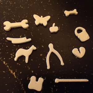 May include: A collection of small, off-white decorative shapes scattered on a dark surface. The shapes include a butterfly, dog bone, apple, dog, wishbone, heart, and other abstract forms. These could be used for crafts or embellishments.