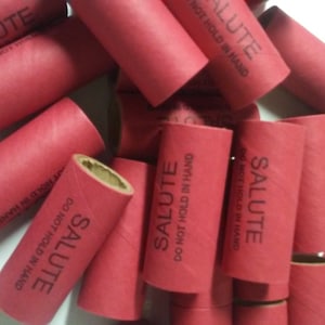 May include: A collection of red cylindrical tubes with the word "SALUTE" printed on them in white. The text "DO NOT HOLD IN HAND" is also printed on the tubes. The tubes are made of cardboard.