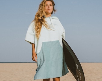 Op de afbeelding: Een vrouw draagt een lichtblauwe poncho met capuchon met een witte top en een zwart surfplank onder haar arm. Ze loopt op een zandstrand.