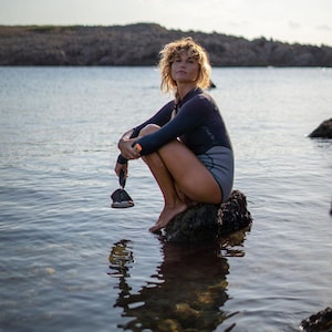 K&ouml;nnte beinhalten: Eine Frau in einem schwarzen Neoprenanzug sitzt auf einem Felsen im Wasser. Sie h&auml;lt ein schwarzes Paddel in ihren H&auml;nden. Das Wasser ist ruhig und der Himmel ist blau.