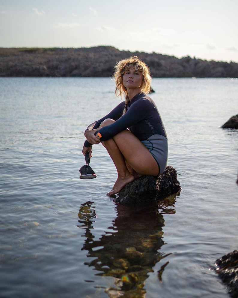 K&ouml;nnte beinhalten: Eine Frau in einem schwarzen Neoprenanzug sitzt auf einem Felsen im Wasser. Sie h&auml;lt ein schwarzes Paddel in ihren H&auml;nden. Das Wasser ist ruhig und der Himmel ist blau.