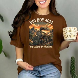May include: A brown t-shirt with a graphic of a steam train, the text "BIG BOY 4014" and "THE LEGEND OF THE RAILS". The person is holding a white mug with gold polka dots and the text "Crate On and Coffee!".