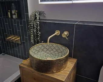 May include: A round vessel sink with a dark exterior and a white interior featuring a repeating black fan-shaped pattern. The sink is mounted on a wooden vanity cabinet and paired with a wall-mounted gold faucet and a matching circular handle.