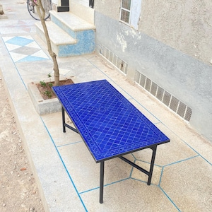 May include: A rectangular blue tile-top table with a black metal frame. The table's surface features a diamond pattern. The table is outdoors, set on a concrete surface with blue trim. The table is approximately 55 inches long and 30 inches wide.