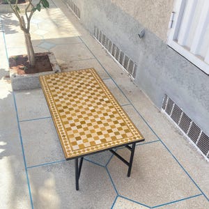 Puede incluir: Mesa rectangular de exterior con tablero a cuadros en tonos beige y marrón. La mesa tiene estructura y patas de metal negro. La mesa mide aproximadamente 152 cm de largo y 76 cm de ancho.