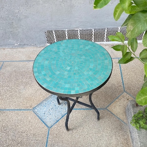 May include: A round outdoor side table with a mosaic tile top in shades of turquoise. The table has a black metal frame and legs. The table is approximately 20 inches in diameter.