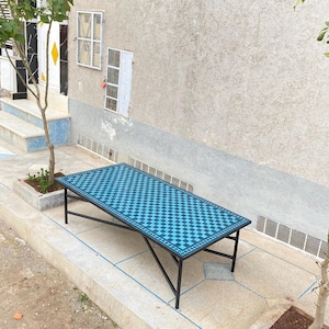 May include: A rectangular outdoor table with a blue and black checkered pattern top. The table has a black metal frame and is set on a concrete patio. The table is approximately 60 inches long.
